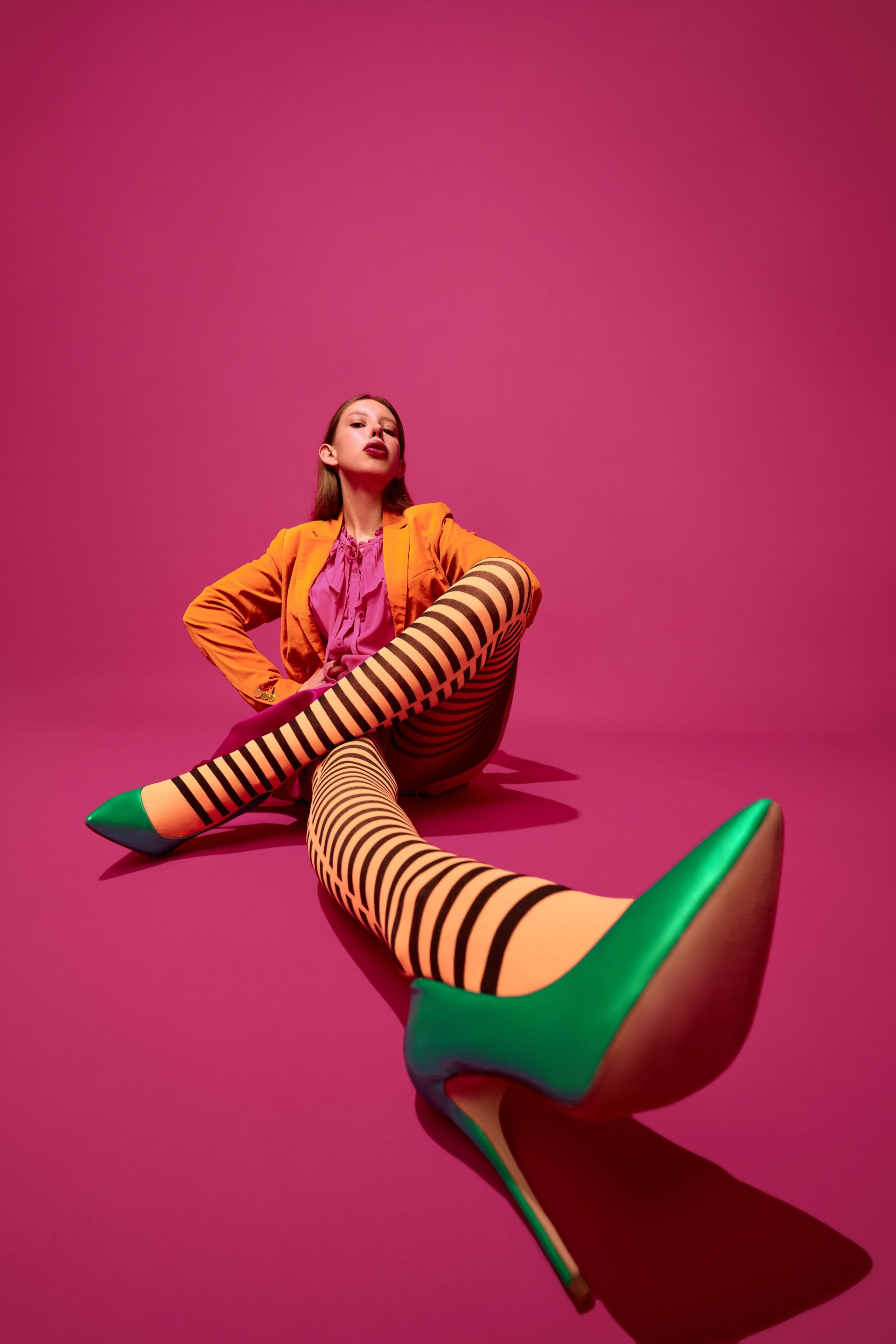 Kreatives Ganzkörperporträt eines ausdrucksstarken jungen Mädchens, Dame in ungewöhnlichen, bunten orange gestreiften Strumpfhosen, langem Rock, kurzer Jacke und grünen Absätzen.